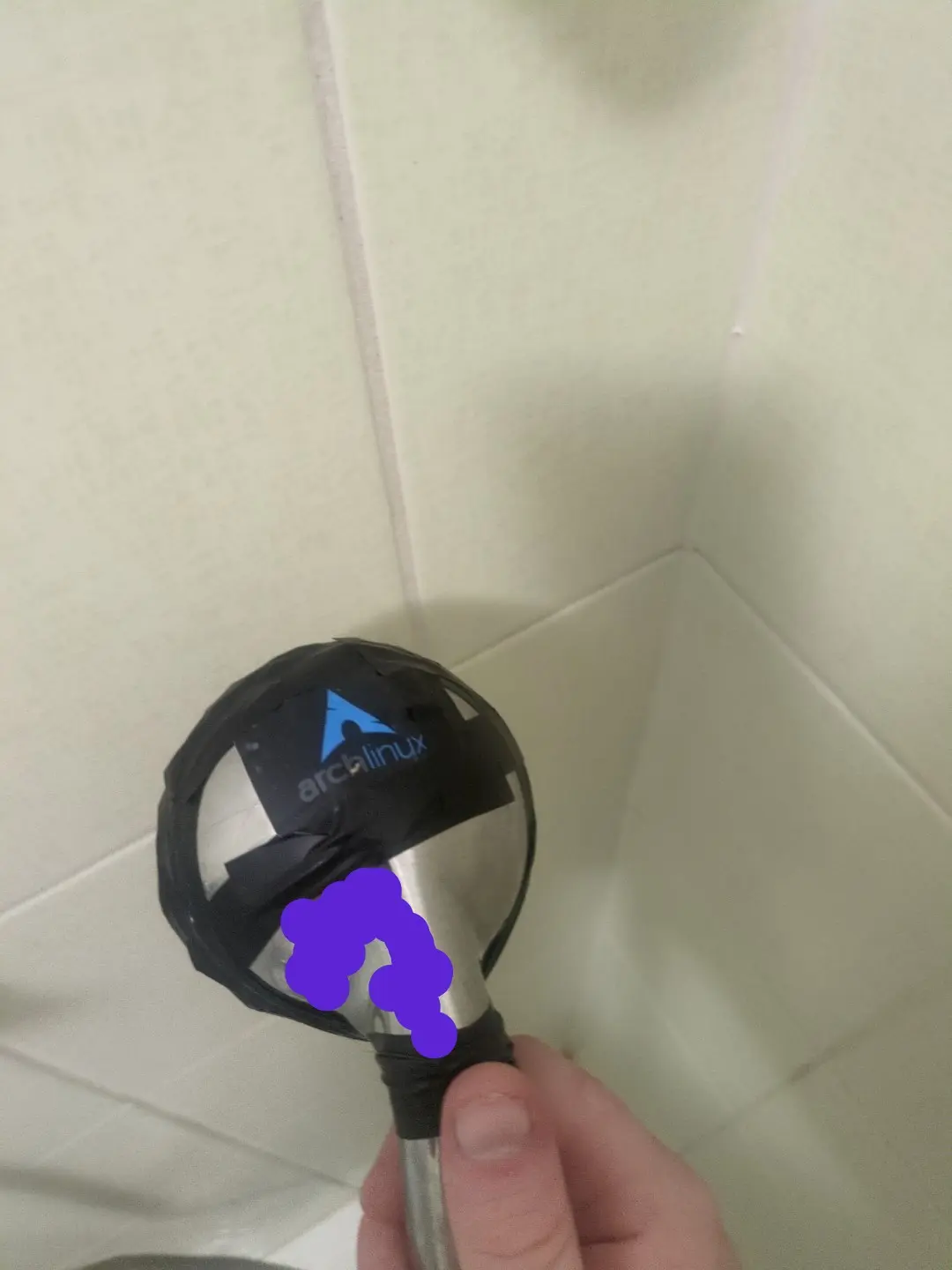 Shower head from the back, arch linux sticker on it and lots of electrical tape. Purple reflection censor.